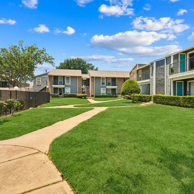 A well-maintained apartment complex with lush green lawns and walking paths. The buildings are two-storied and feature balconies. The skies are bright with fluffy clouds overhead.