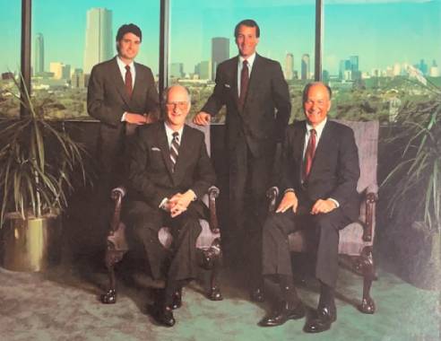 Four business professionals posing in an office setting with a city skyline in the background.