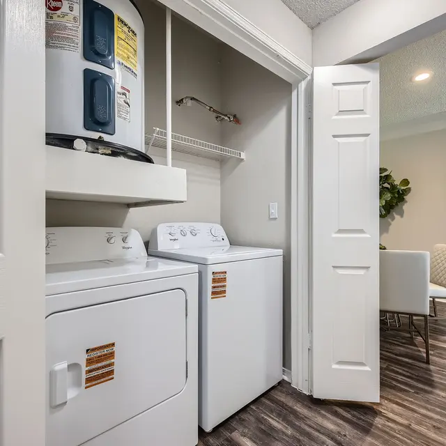 A compact laundry room featuring a stacked washer and dryer with white doors, and a glimpse of a modern living area beyond through an open doorway.