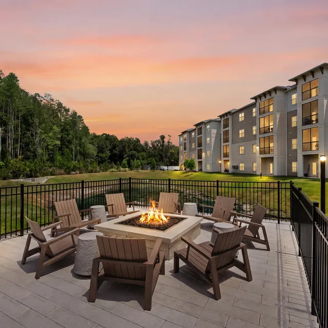 Outdoor Firepit Area at Sunset An outdoor firepit area with wooden chairs surrounded by greenery and a residential building in the background during dusk.