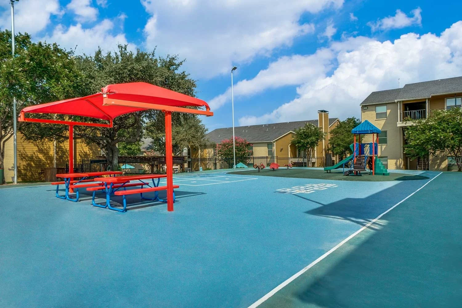 Outdoor playground area featuring a colorful play structure, picnic tables with a red canopy, and surrounding greenery.