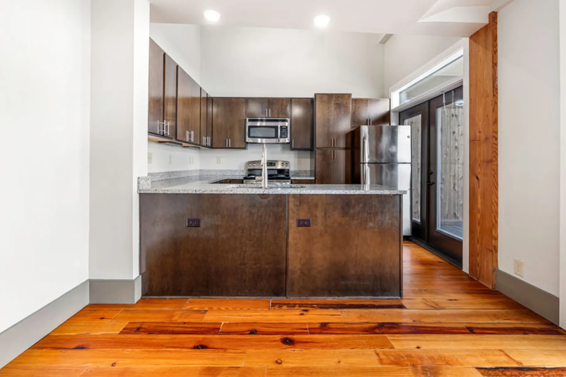 A modern kitchen featuring dark wood cabinetry, granite countertops, and stainless steel appliances. The floor is made of polished hardwood.