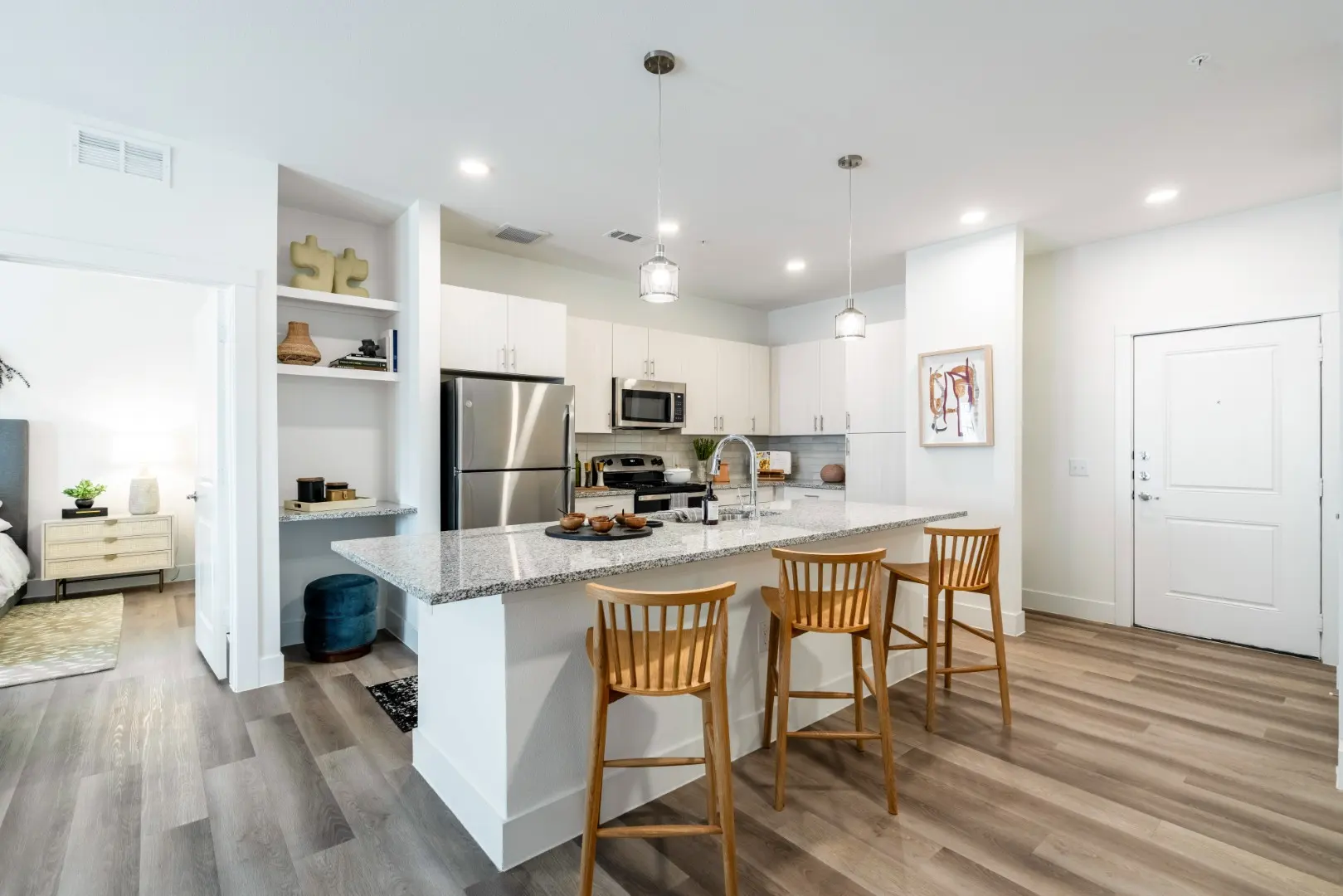 A modern kitchen featuring a spacious island with four wooden barstools, stainless steel appliances, and a cozy ambiance. The kitchen has a white cabinetry and a light-colored countertop. Visible is an open layout leading to a bedroom area.